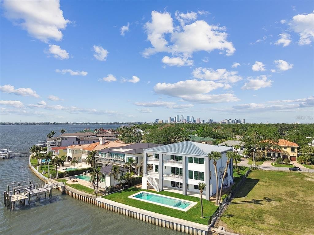 This aerial view showcases a luxurious waterfront property featuring a modern, multi-story home with a pool and meticulously maintained lawn. The property includes a private dock extending into the water, complemented by neighboring upscale residences and a distant city skyline. The overall impression is one of exclusivity and high-end coastal living.