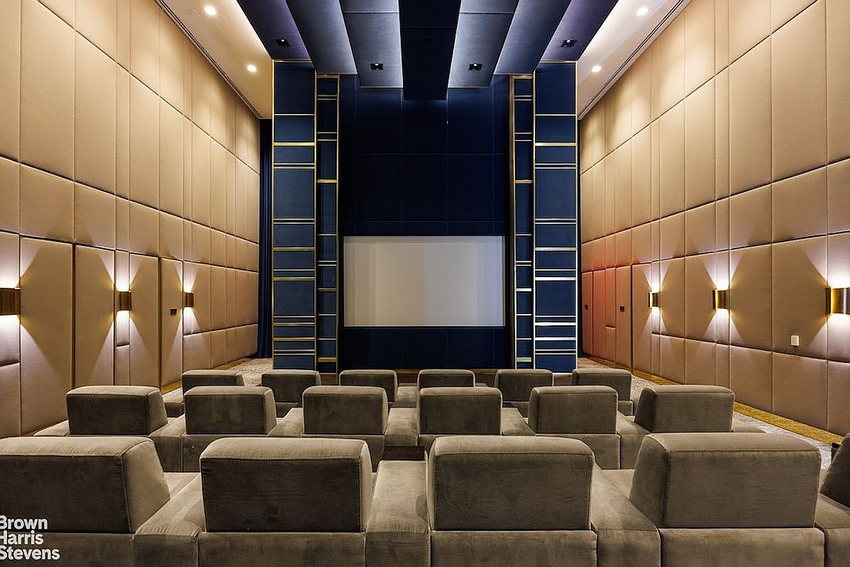 This image showcases a sophisticated private home theater featuring tiered rows of plush, neutral-toned seating facing a large projection screen. The room is framed by tall, padded wall panels in a warm tan hue, accented by symmetrical wall sconces and a striking central feature wall in deep blue with gold-toned vertical trim. The perspective is centered from the back of the room, emphasizing the symmetrical layout and the luxurious, cinematic atmosphere of the space.