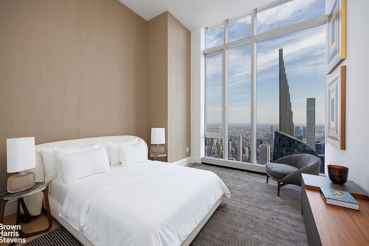 This modern primary bedroom features a minimalist aesthetic with a neutral color palette and a plush bed centered against a textured accent wall. The room is defined by expansive floor-to-ceiling windows that provide a dramatic, high-altitude view of the city skyline, including a prominent skyscraper. A sleek armchair sits by the window, and a low-profile wooden console table adds a touch of warmth to the sophisticated, high-rise space.