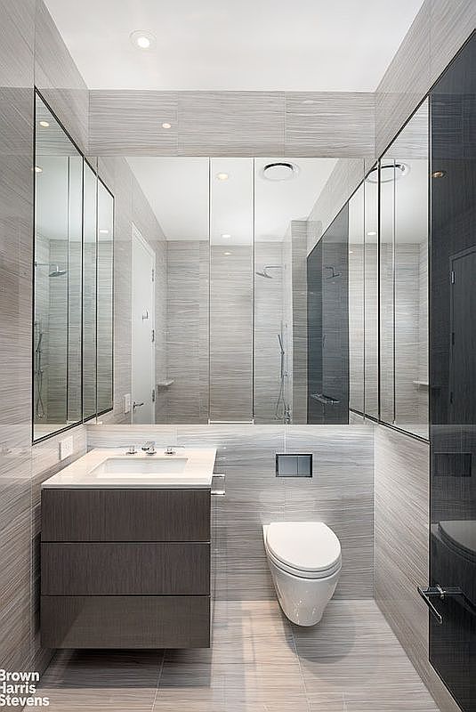 This modern, compact bathroom features a sleek floating vanity with a rectangular sink and a wall-mounted toilet. The space is finished with horizontal-patterned light gray tiles that cover the walls and floor, creating a cohesive and contemporary aesthetic. A large, multi-paneled mirrored medicine cabinet spans the width of the vanity, reflecting the shower area and enhancing the sense of space in the room.