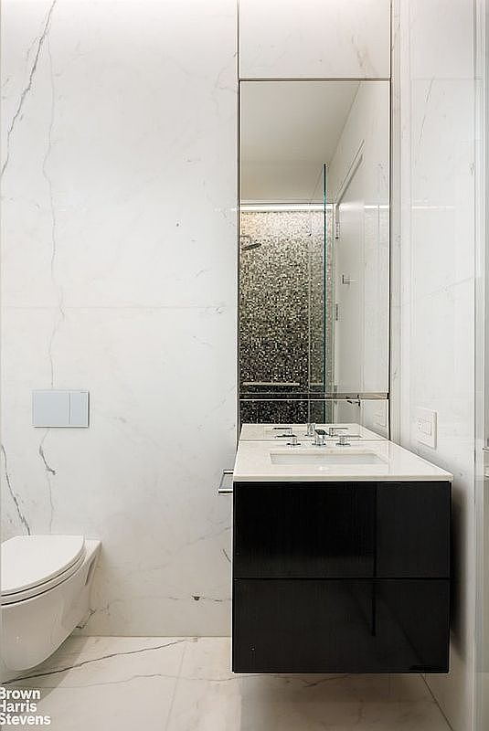 This modern bathroom features a sleek, wall-mounted black vanity with a white countertop and a rectangular sink. The walls and floor are covered in large-format light-colored stone tiles with subtle veining, creating a clean and minimalist aesthetic. A tall, frameless mirror sits above the vanity, reflecting the shower area which is enclosed by glass and finished with textured, dark-toned wall tiles.