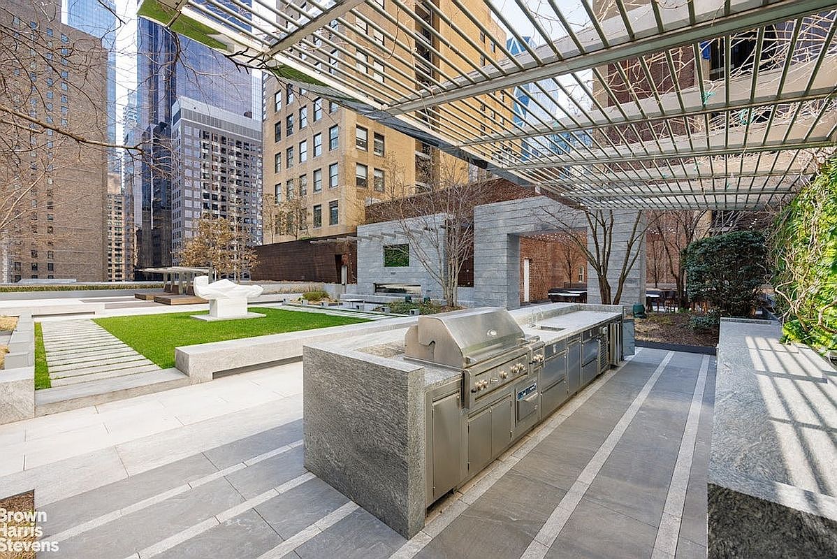 This expansive urban terrace features a sophisticated outdoor kitchen equipped with a stainless steel grill and stone-clad prep area, positioned beneath a modern metal pergola. The space includes a manicured lawn with a sculptural art piece, stone pathways, and a stone-framed architectural structure in the background. Surrounded by high-rise city buildings, this terrace offers a luxurious and private outdoor retreat in a metropolitan setting.