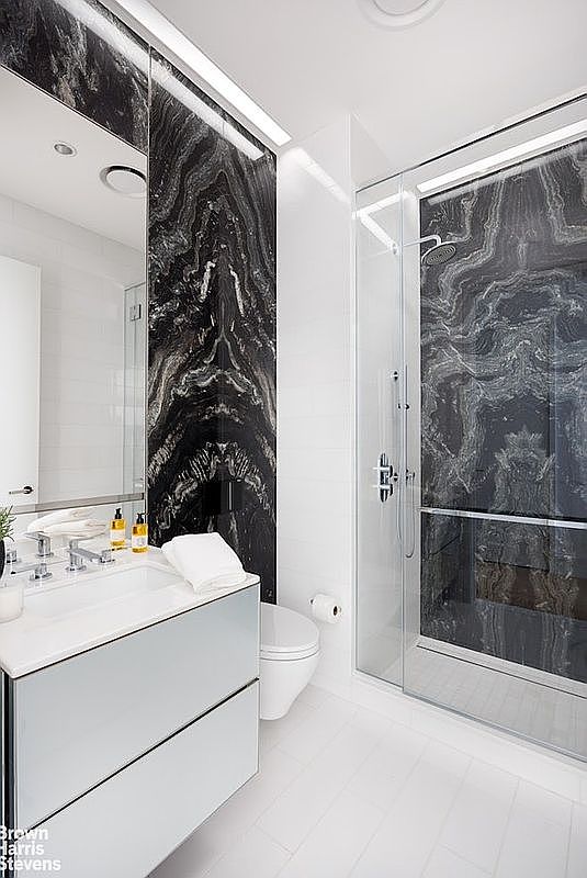 This modern bathroom features a striking contrast between white surfaces and dramatic, dark veined stone panels that accent the vanity wall and the shower enclosure. The space includes a sleek, wall-mounted vanity with a white countertop, a glass-enclosed walk-in shower, and a white toilet. The perspective is a wide-angle shot from the doorway, highlighting the clean lines and high-contrast aesthetic of the contemporary design.