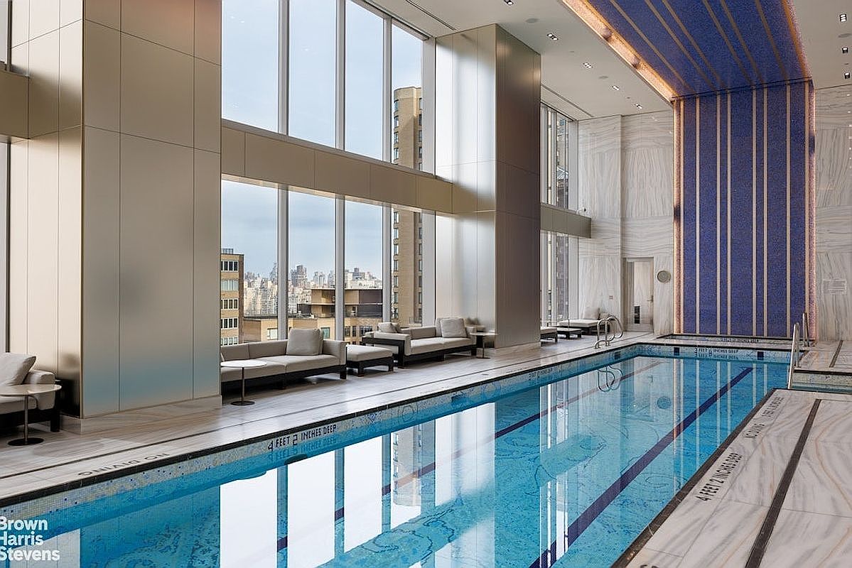 This luxurious indoor swimming pool area features a long lap pool lined with light-colored stone and marked with depth indicators. Large windows span the length of the room, offering expansive city views and flooding the space with natural light, while plush lounge seating is arranged along the perimeter. The room is accented by a striking blue mosaic-tiled wall feature and high ceilings, creating an elegant and serene atmosphere.