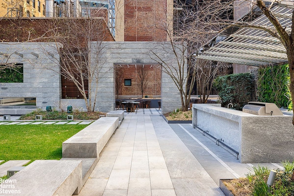 This sophisticated outdoor terrace features a stone-paved walkway leading through a central archway into a secondary seating area. The space is equipped with a built-in stone outdoor kitchen and grill, complemented by a manicured lawn and mature trees. The architectural design blends modern stone elements with natural landscaping, creating a serene and upscale urban retreat.