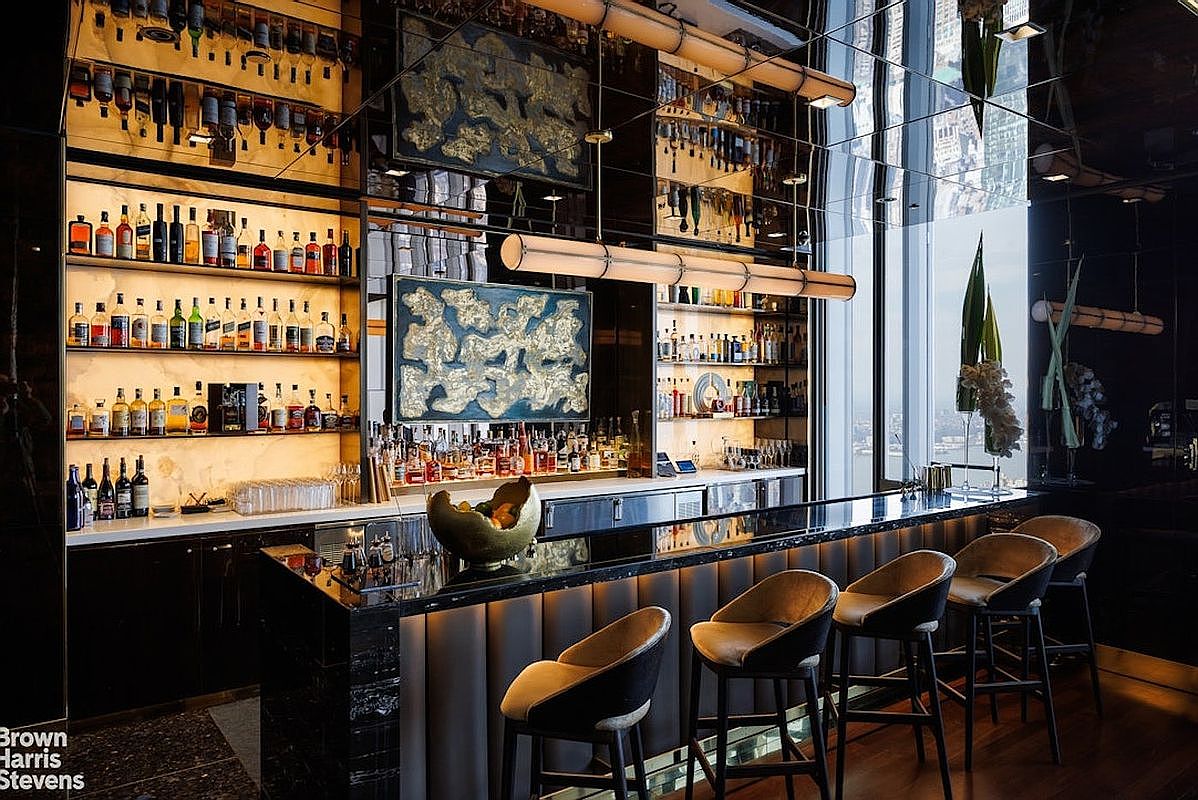 This sophisticated home bar area features a sleek, dark stone-topped counter with integrated bar seating and a row of four modern, upholstered stools. The back bar is elegantly illuminated with warm lighting, showcasing an extensive collection of spirits on multiple glass shelves, accented by abstract artwork and mirrored surfaces. The space offers a refined, upscale atmosphere with a view of the city skyline visible through the large windows to the right.