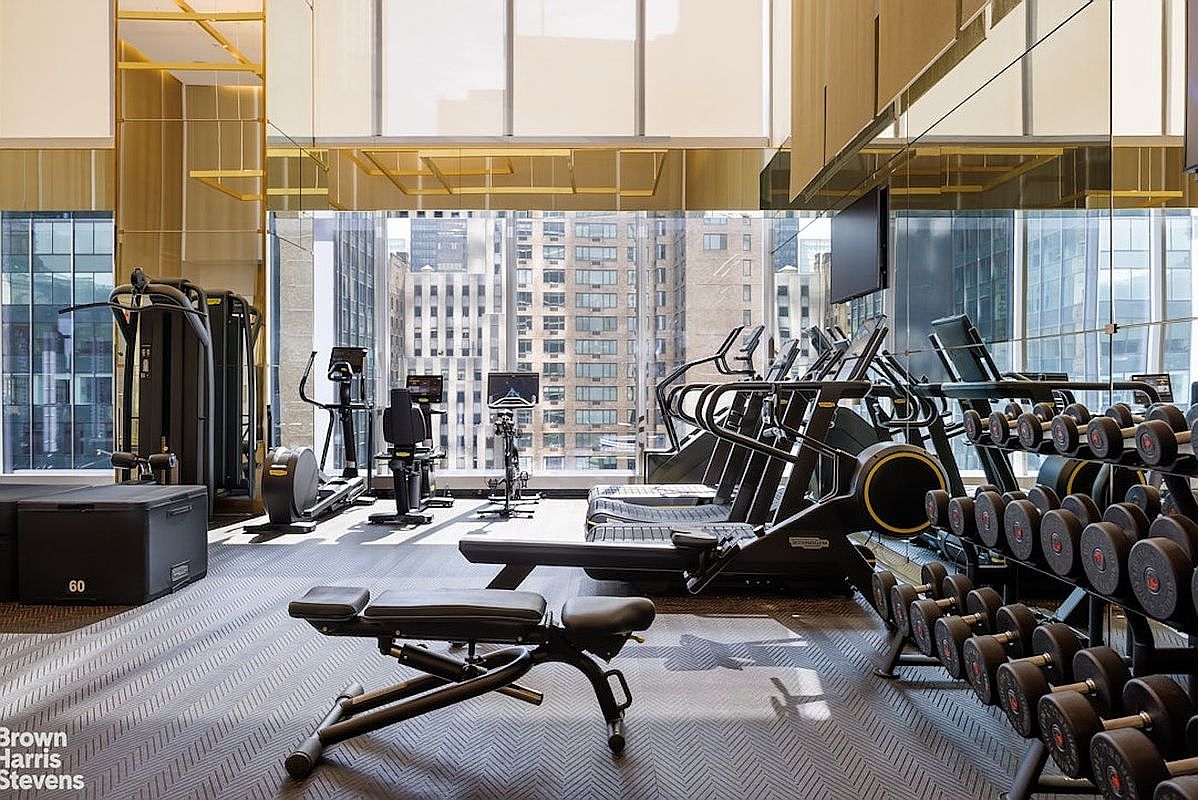 This professional-grade home gym features a sleek, modern aesthetic with high-end fitness equipment, including treadmills, a stationary bike, and a comprehensive rack of dumbbells. The space is defined by its expansive, floor-to-ceiling windows that provide an impressive, elevated view of the surrounding city skyline. The room is finished with a textured, dark-toned floor and mirrored walls that enhance the sense of depth and light within the workout area.