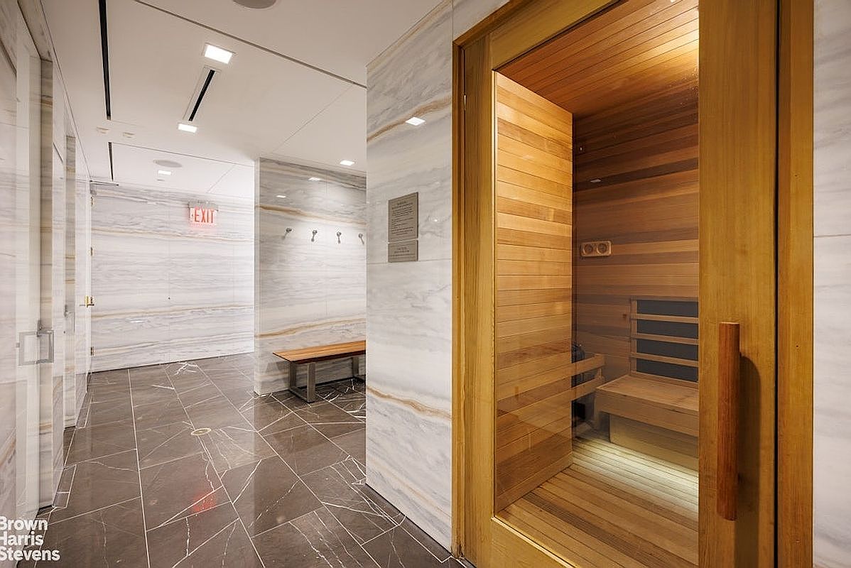 This image showcases a sleek, modern sauna room integrated into a shared facility, featuring warm wood paneling and a glass door. The adjacent hallway is finished with polished stone flooring and light-toned stone wall cladding, creating a clean and upscale aesthetic. A wooden bench and wall-mounted hooks are visible in the hallway, providing a functional space for residents.