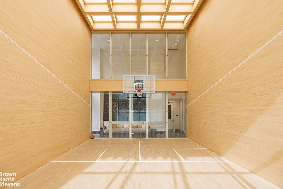This indoor basketball court features light wood paneling on the walls and floor, creating a bright and cohesive aesthetic. A glass wall separates the court from an adjacent lounge area, while a large skylight grid above provides ample natural light. The perspective is centered, looking directly at the basketball hoop mounted on the glass partition.