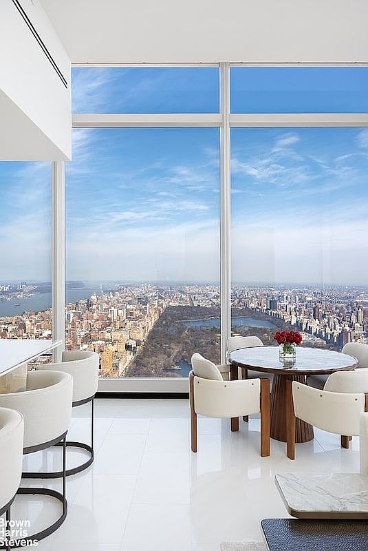 This high-rise dining area features a circular table surrounded by modern, cream-colored upholstered chairs, set against a backdrop of expansive floor-to-ceiling windows. The space offers a breathtaking, unobstructed panoramic view of Central Park and the surrounding New York City skyline. The interior is characterized by a minimalist aesthetic, featuring polished white flooring and a sleek, contemporary design that emphasizes the dramatic urban vista.