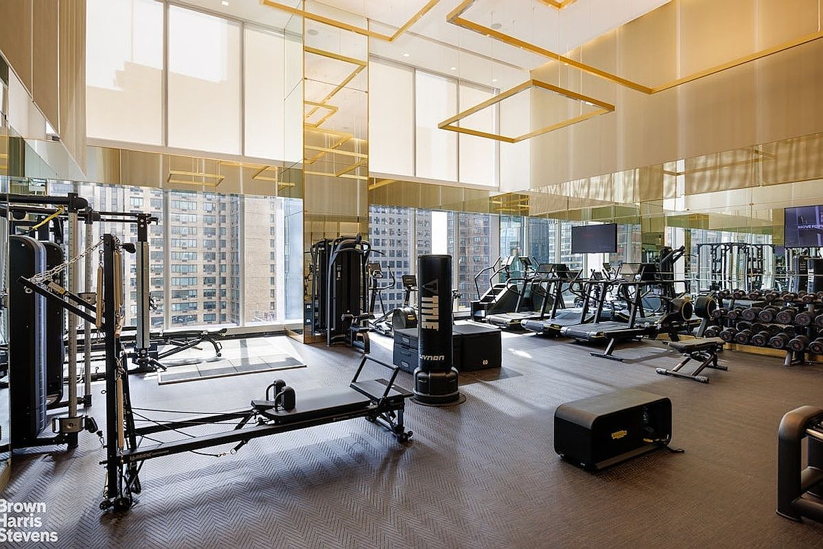 This high-end, professional-grade home gym features a spacious layout with a variety of fitness equipment, including a Pilates reformer, a boxing heavy bag, and multiple cardio machines. The room is defined by its expansive floor-to-ceiling windows that offer panoramic city views, complemented by mirrored walls and elegant gold-toned geometric ceiling accents. The overall aesthetic is sophisticated and modern, creating an upscale environment for exercise.