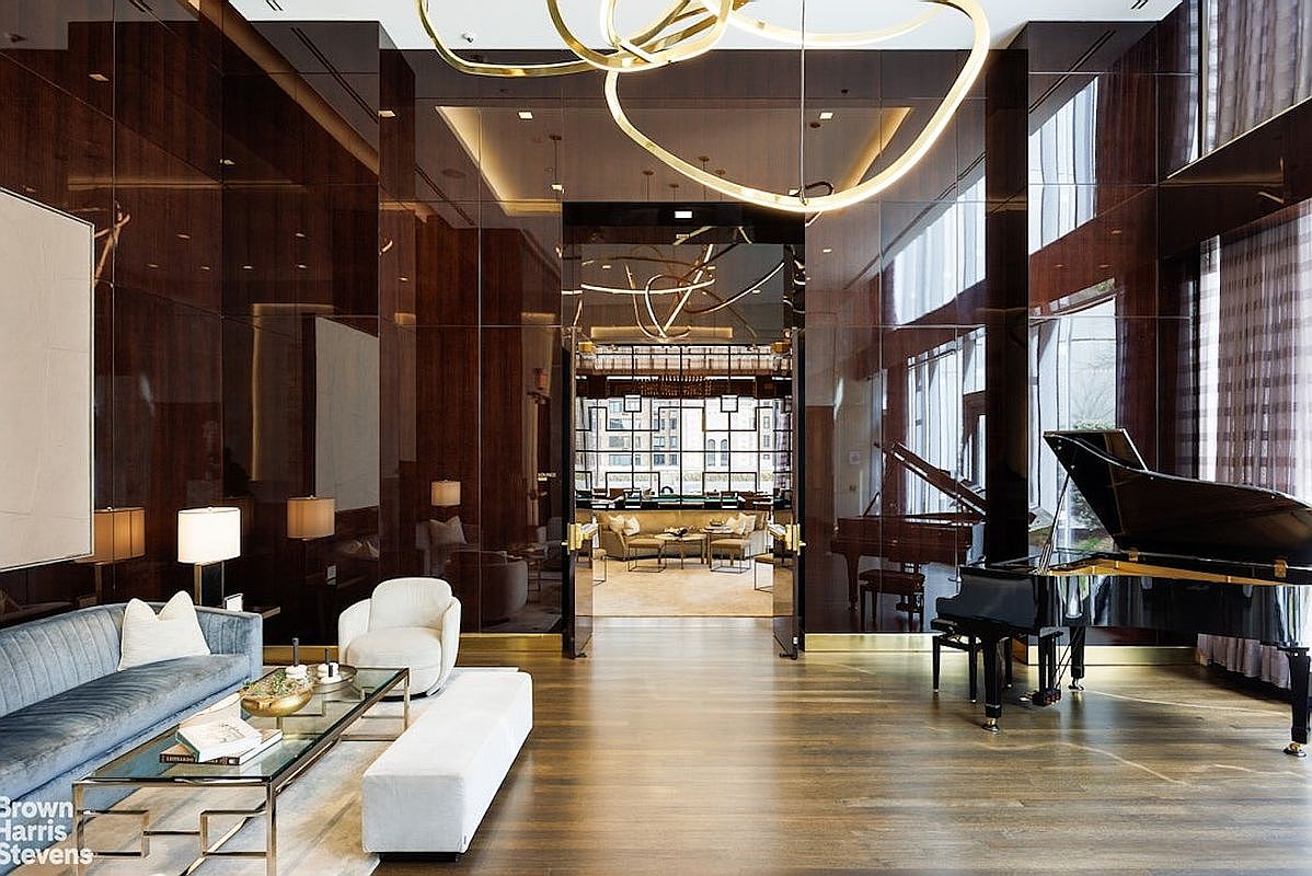 This sophisticated, high-end lounge area features dark, reflective wall paneling and polished wood flooring, creating a luxurious and moody atmosphere. A grand piano sits prominently on the right, while a plush blue sofa and white accent chair provide comfortable seating in the foreground. The space is illuminated by a striking, sculptural gold light fixture and leads through a central doorway into an adjacent, brightly lit seating area.