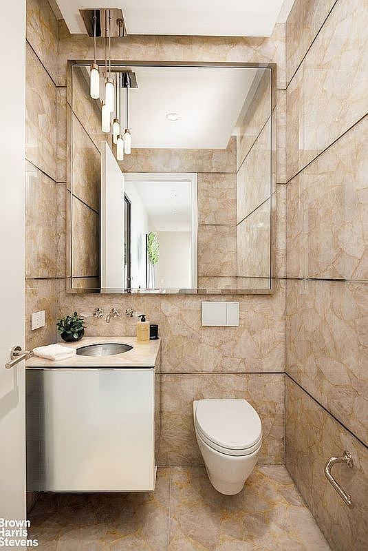 This compact powder room features striking, light-toned stone tile cladding on the walls and floor, creating a cohesive and luxurious aesthetic. A sleek, metallic-finished vanity with an integrated sink sits beneath a large, frameless mirror, which is illuminated by a modern, multi-pendant light fixture. The space is completed by a wall-mounted toilet and minimalist hardware, offering a sophisticated and contemporary design.