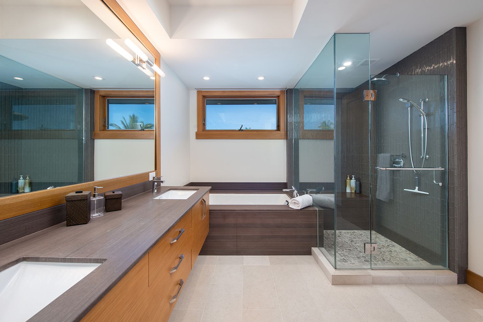 This is a well-lit primary bathroom featuring a double vanity with modern fixtures and a large mirror. A soaking tub is nestled beneath a window, and a glass-enclosed shower with pebble flooring adds a luxurious touch. The color palette is neutral, creating a serene and spa-like atmosphere.