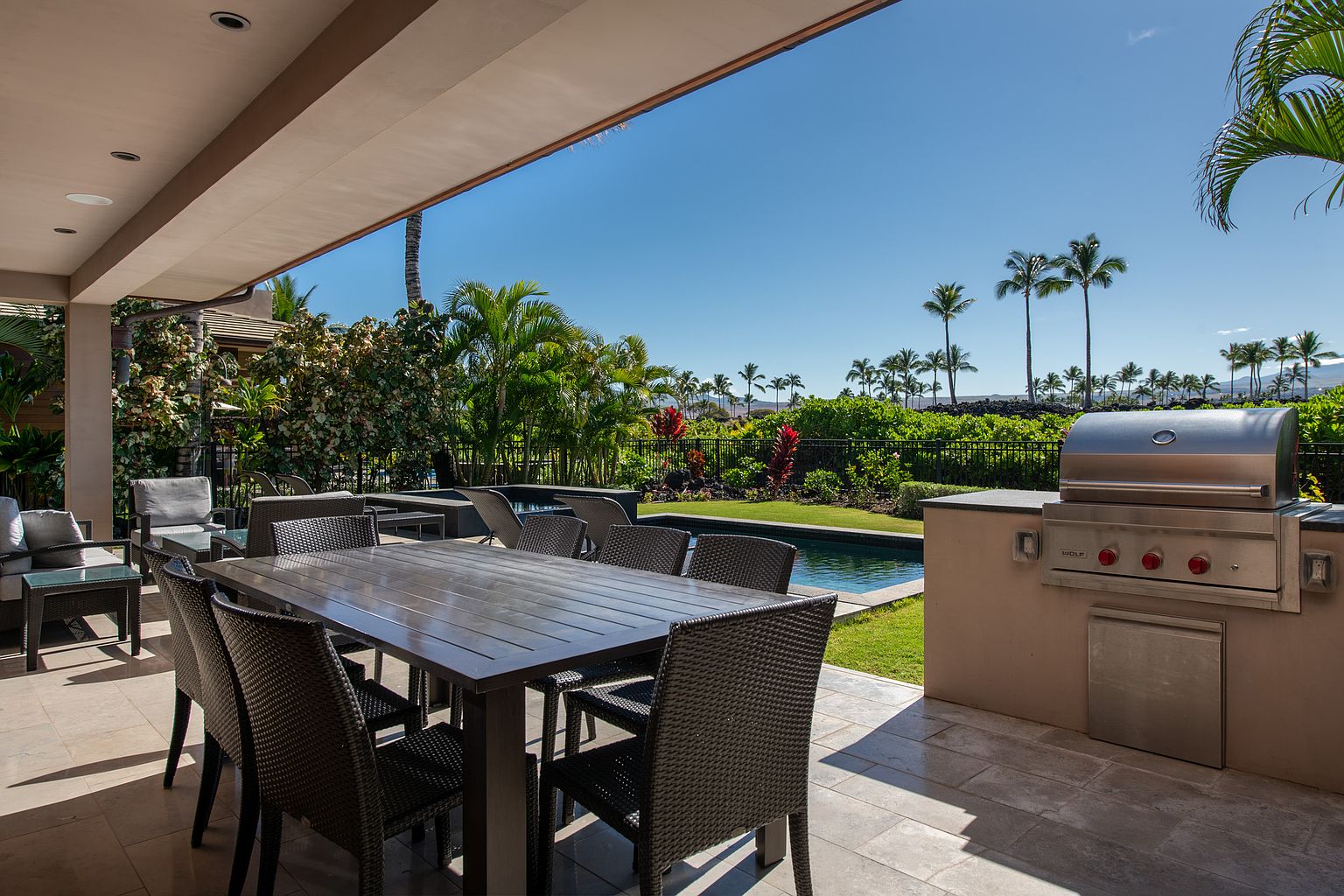 This is a view of an outdoor patio area featuring a large dining table with woven chairs, a built-in grill, and a swimming pool in the background. The patio is covered by an overhang, providing shade, and the landscaping includes palm trees and lush greenery. The scene suggests a luxurious outdoor living space perfect for entertaining.