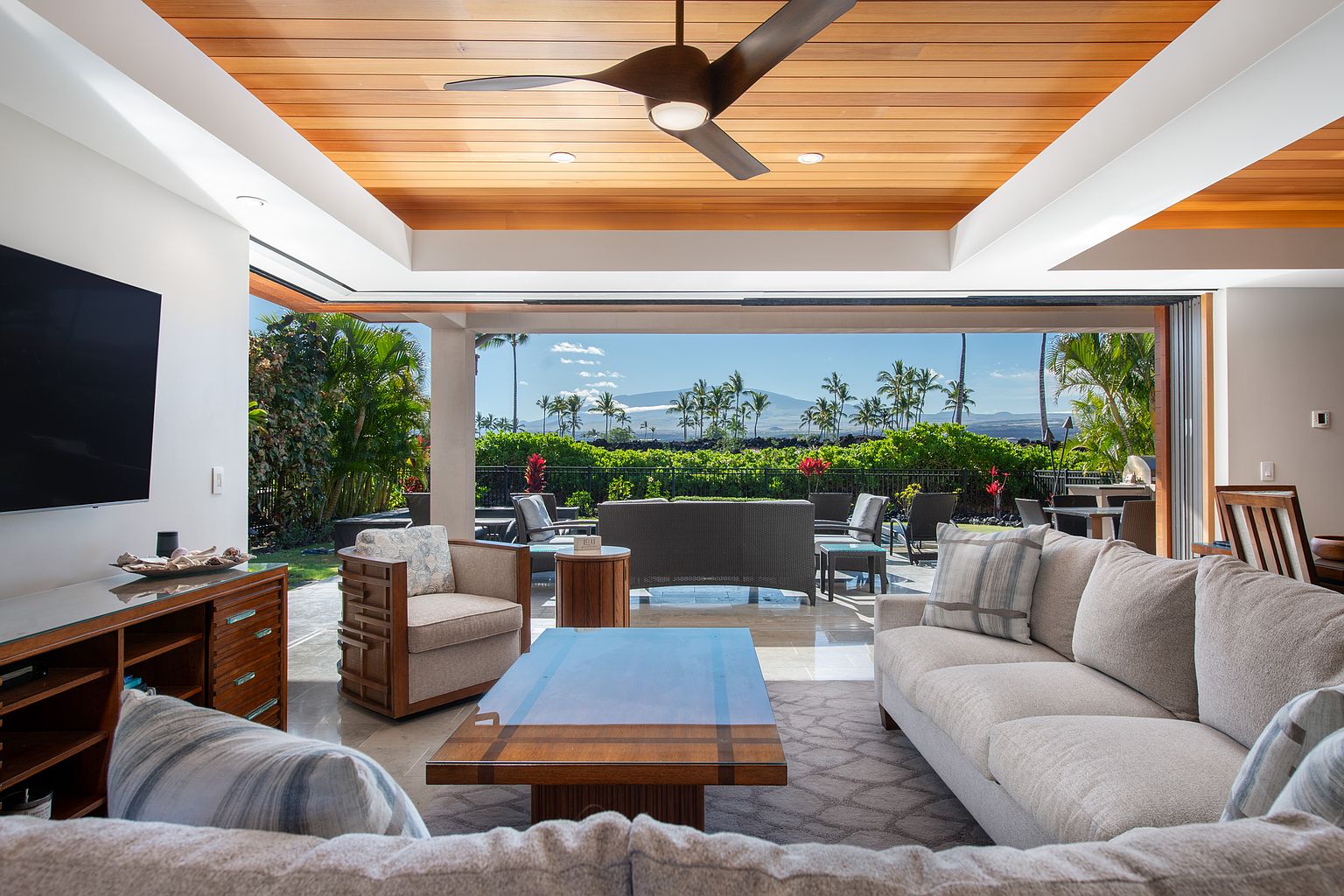 This is a spacious living room that seamlessly blends indoor and outdoor living with large sliding glass doors opening to a patio. The room features comfortable seating arrangements, including a sofa and armchairs, arranged around a coffee table. A ceiling fan adds to the comfort, while the view of the lush greenery and distant landscape enhances the appeal of this inviting space.