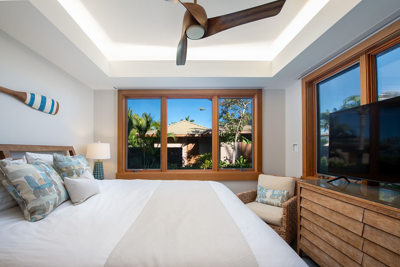 This is a primary bedroom featuring a king-size bed with white linens and decorative pillows. The room has a tray ceiling with recessed lighting and a ceiling fan. Large windows provide natural light and views of the exterior landscape, while a wooden dresser with a television and a wicker chair add to the room's comfort and style.