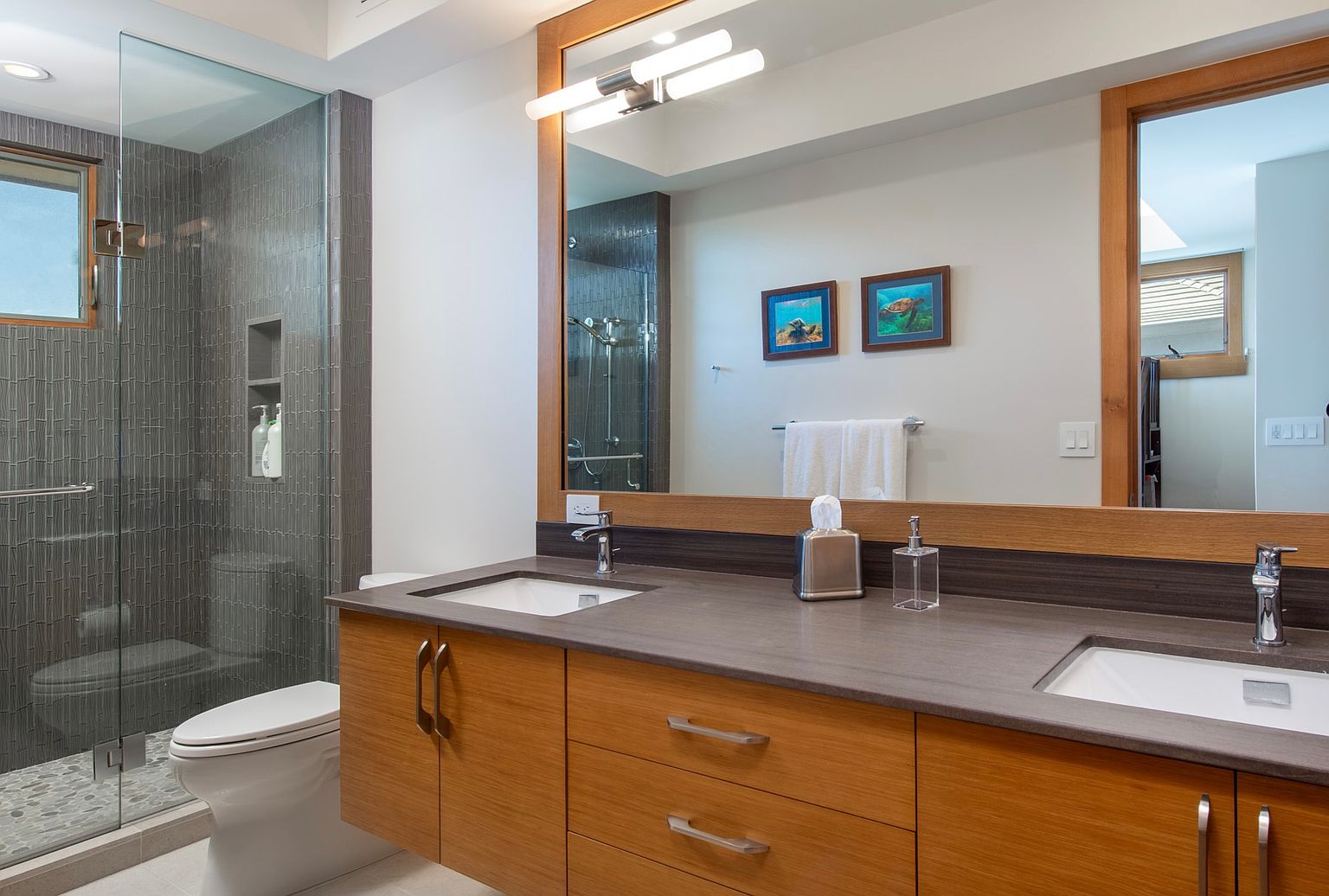 This is a well-lit primary bathroom featuring a double vanity with a dark countertop and light wood cabinetry. A large mirror reflects the vanity area and part of the shower, which has gray tiled walls and a glass enclosure. The overall impression is clean, modern, and functional.