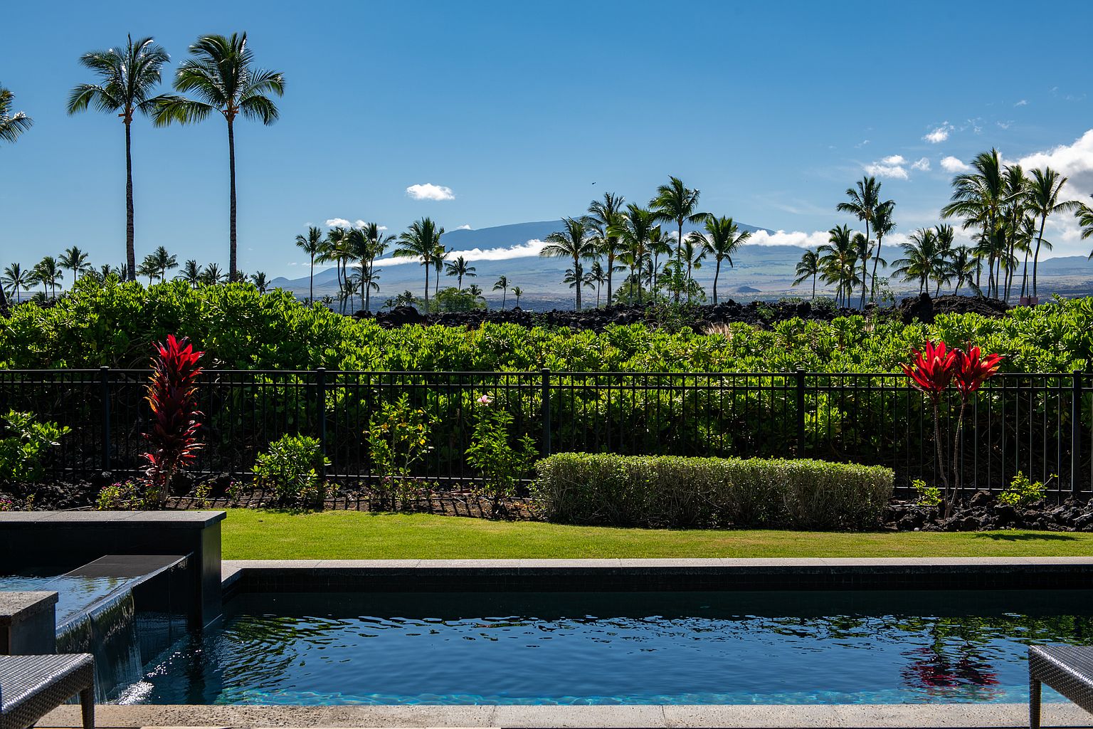 This image showcases a luxurious pool area with a serene view. The pool's dark blue water reflects the surrounding landscape, which includes lush greenery, palm trees, and a distant mountain range under a clear blue sky. A sleek, modern waterfall feature adds to the upscale ambiance, making it an inviting outdoor space.