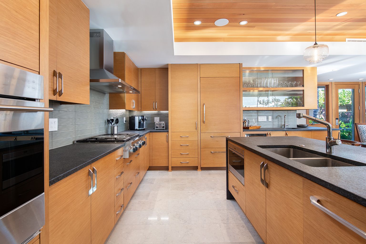 This is a well-lit, modern kitchen featuring light wood cabinetry and dark countertops. Stainless steel appliances are integrated throughout, including a double oven and a range with a stainless steel hood. The kitchen island provides additional counter space and storage, while the light-colored tile flooring adds to the bright and airy feel of the space.