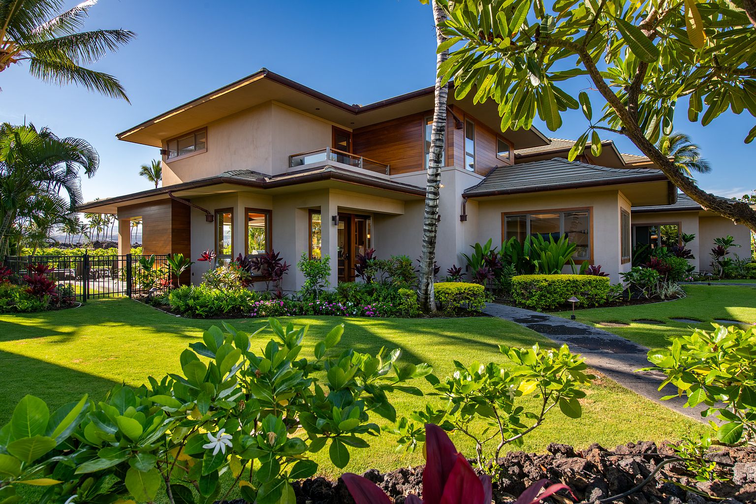 This is a front exterior view of a luxurious two-story home. The house features a combination of stucco and wood siding, with a well-manicured lawn and lush landscaping. The architectural style is modern with clean lines and a warm, inviting color palette, suggesting a high-end property with excellent curb appeal.