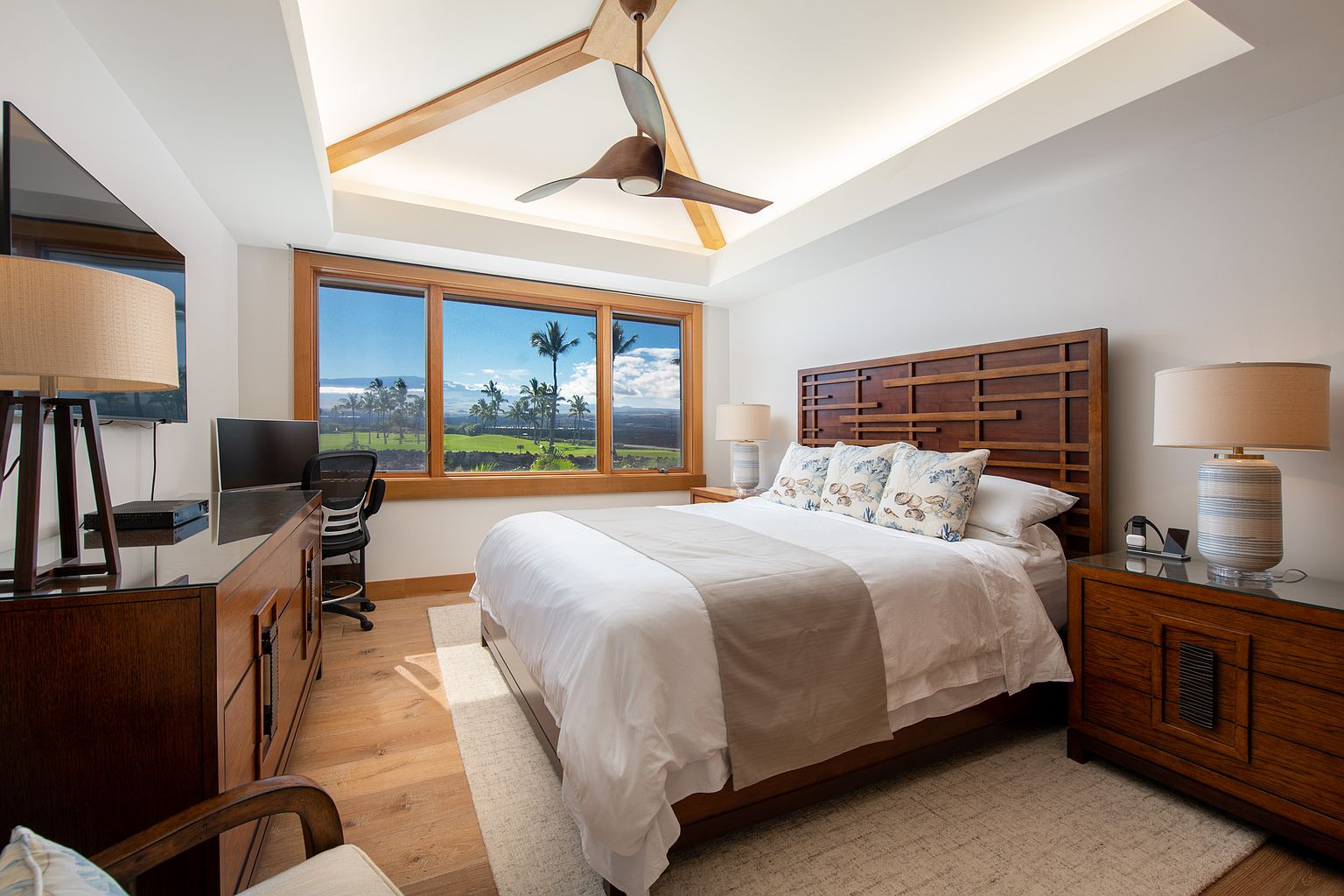This primary bedroom features a king-size bed with a decorative headboard and neutral-toned bedding. A large window offers a view of a lush green landscape with palm trees. The room is furnished with wooden dressers and lamps, and a ceiling fan adds to the comfort of the space, creating a serene and inviting atmosphere.