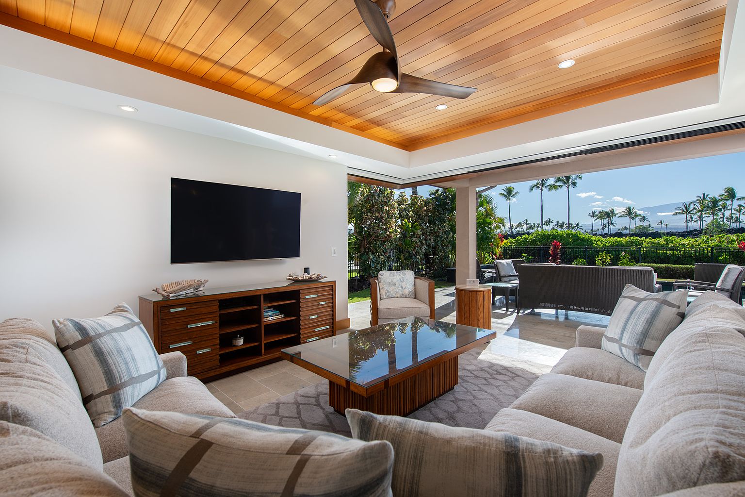 This is a bright and airy living room that seamlessly blends indoor and outdoor living. The room features a large sectional sofa with decorative pillows, a wooden entertainment center with a flat-screen TV, and a glass-topped coffee table. The wooden ceiling adds warmth, while the open patio doors reveal a lush tropical landscape, creating a relaxing and inviting atmosphere.