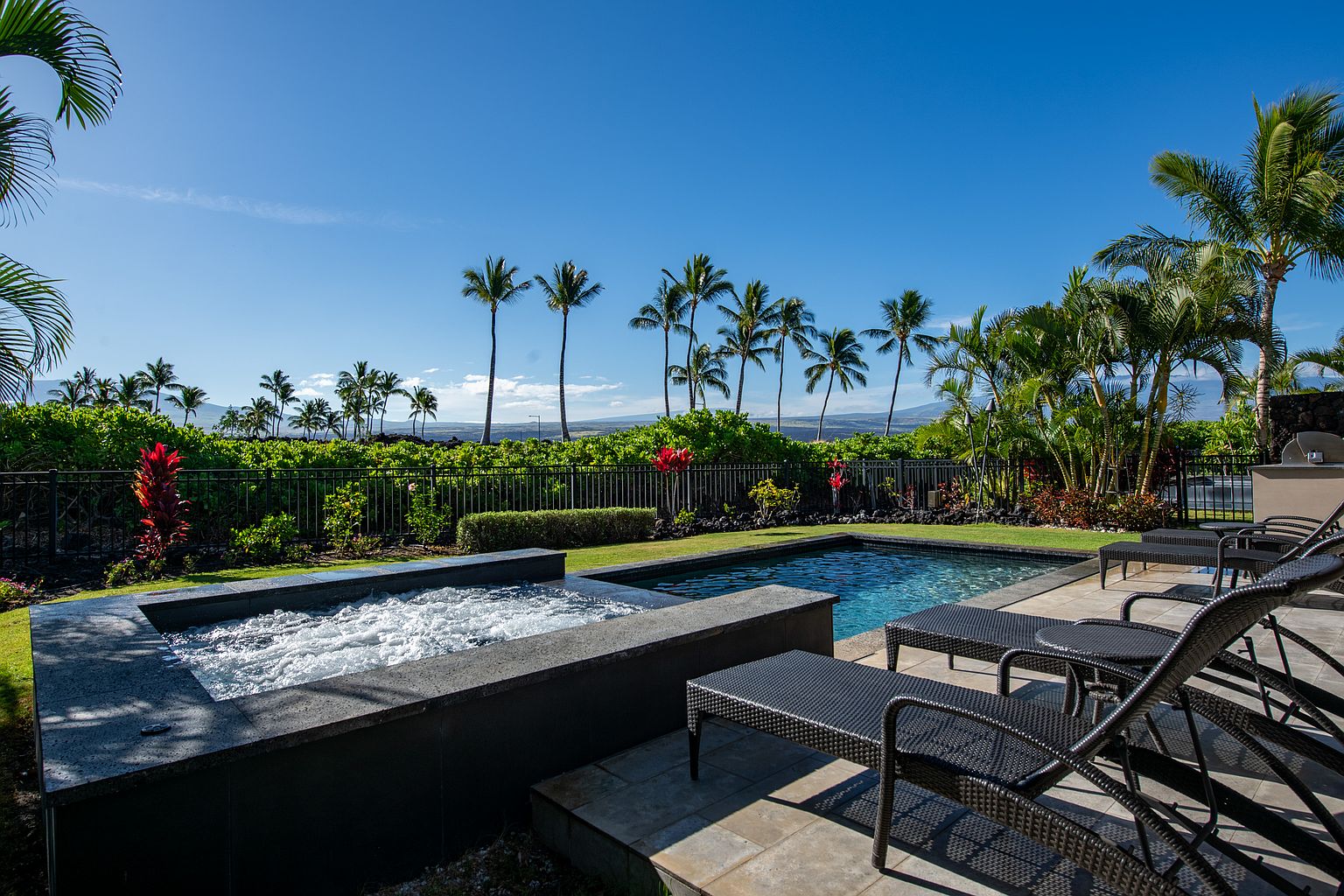 This image showcases a luxurious backyard pool and spa area, perfect for relaxation and entertainment. The scene features a modern spa with bubbling water, a sleek pool, and comfortable lounge chairs arranged on a tiled patio. Lush tropical landscaping and palm trees create a serene and private oasis, enhancing the property's appeal.