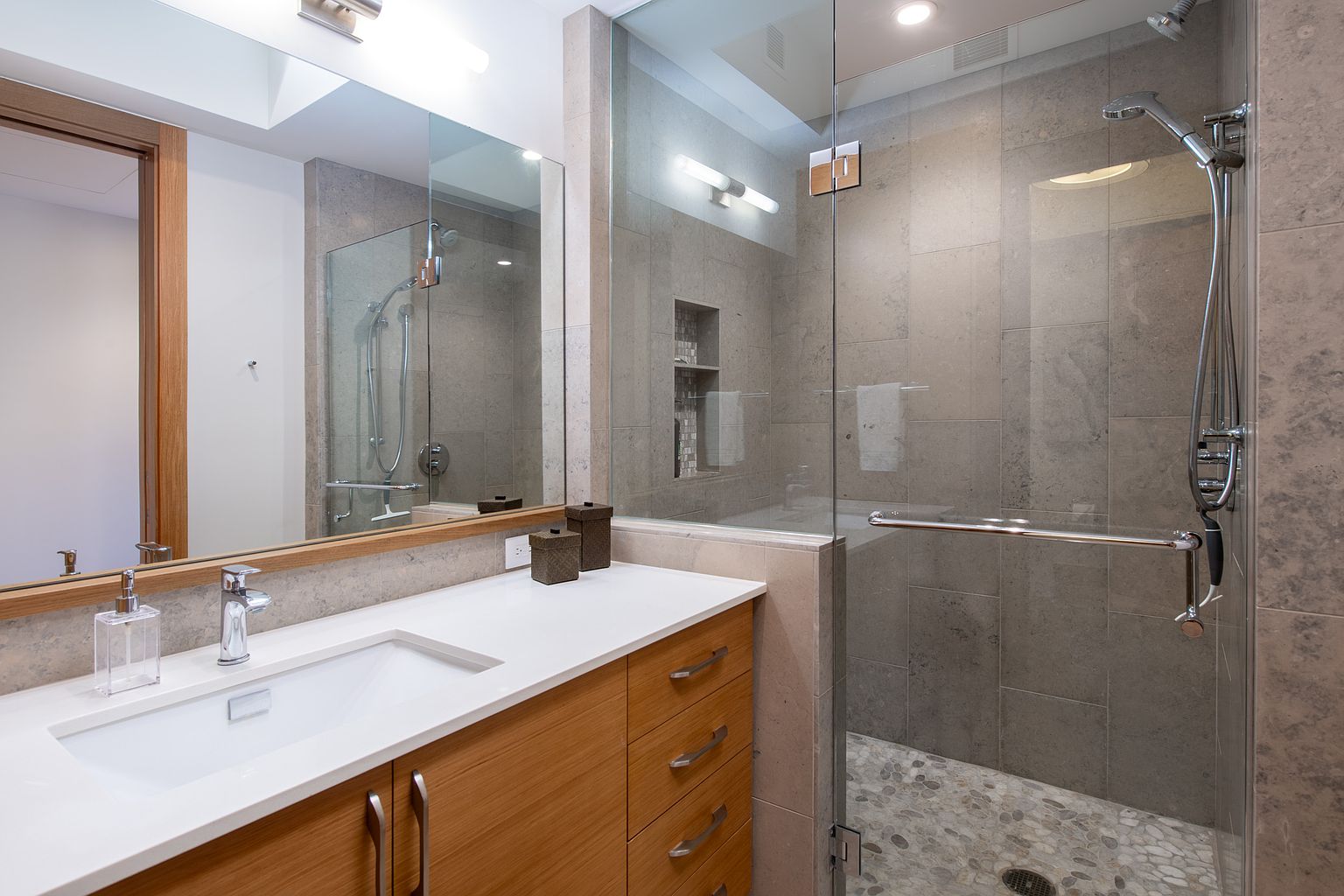 This is a well-lit primary bathroom featuring a modern design with a wood-finish vanity, a white countertop, and a rectangular sink. A large mirror reflects the shower area, which is enclosed with glass and has gray tiling and a pebble floor. The overall impression is clean, contemporary, and luxurious.