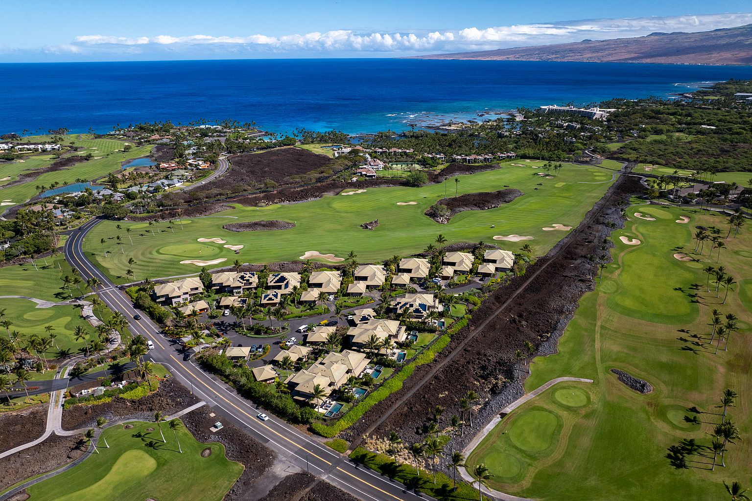 This aerial shot showcases a luxurious residential area nestled within a golf course community. The homes feature tan roofs and some have private pools, surrounded by lush greenery and palm trees. The scene is framed by the blue ocean and a distant mountain range, creating a serene and upscale atmosphere.