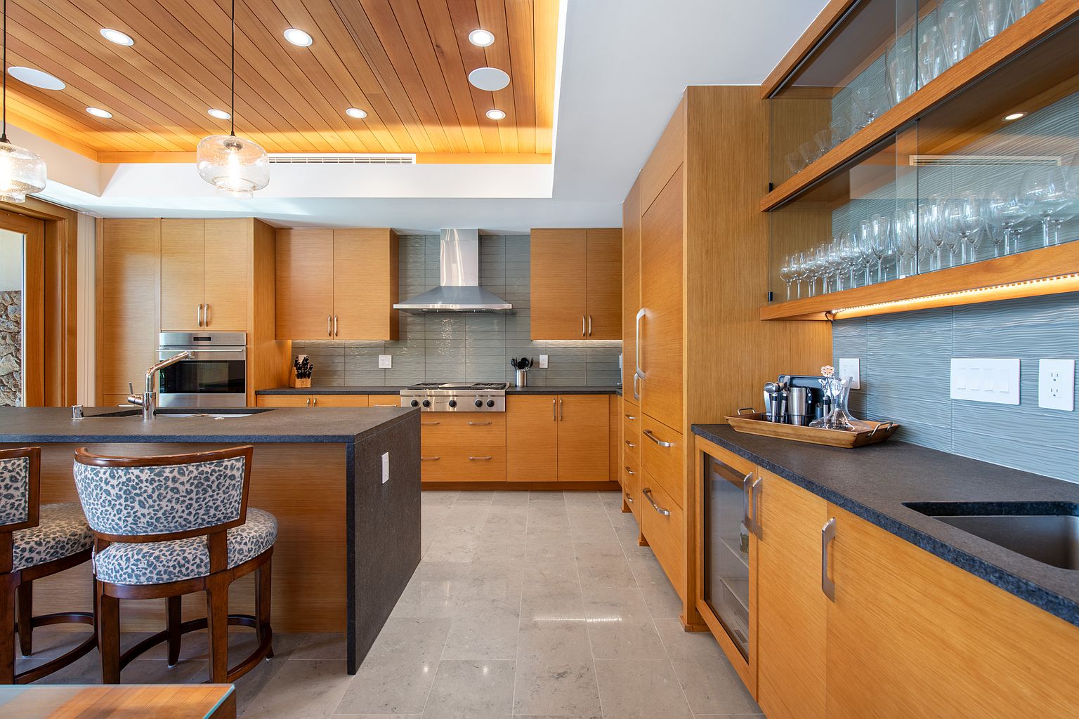 This is a well-lit, modern kitchen featuring light wood cabinetry, dark countertops, and stainless steel appliances. The kitchen island provides additional workspace and seating, while the open shelving displays glassware. The overall design is sleek and functional, creating a welcoming space for cooking and entertaining.