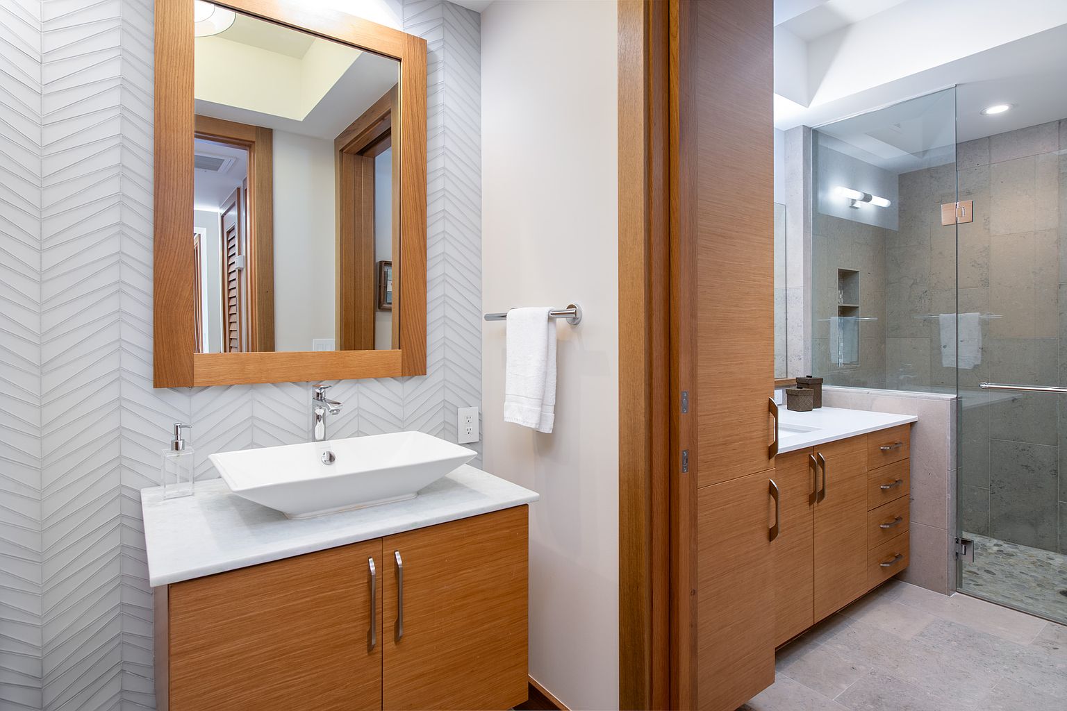 This is a well-lit bathroom featuring a modern vanity with a white countertop and a vessel sink. The walls are adorned with a subtle chevron pattern, and a wooden-framed mirror hangs above the sink. A glass-enclosed shower is visible in the background, adding to the contemporary aesthetic of the space.