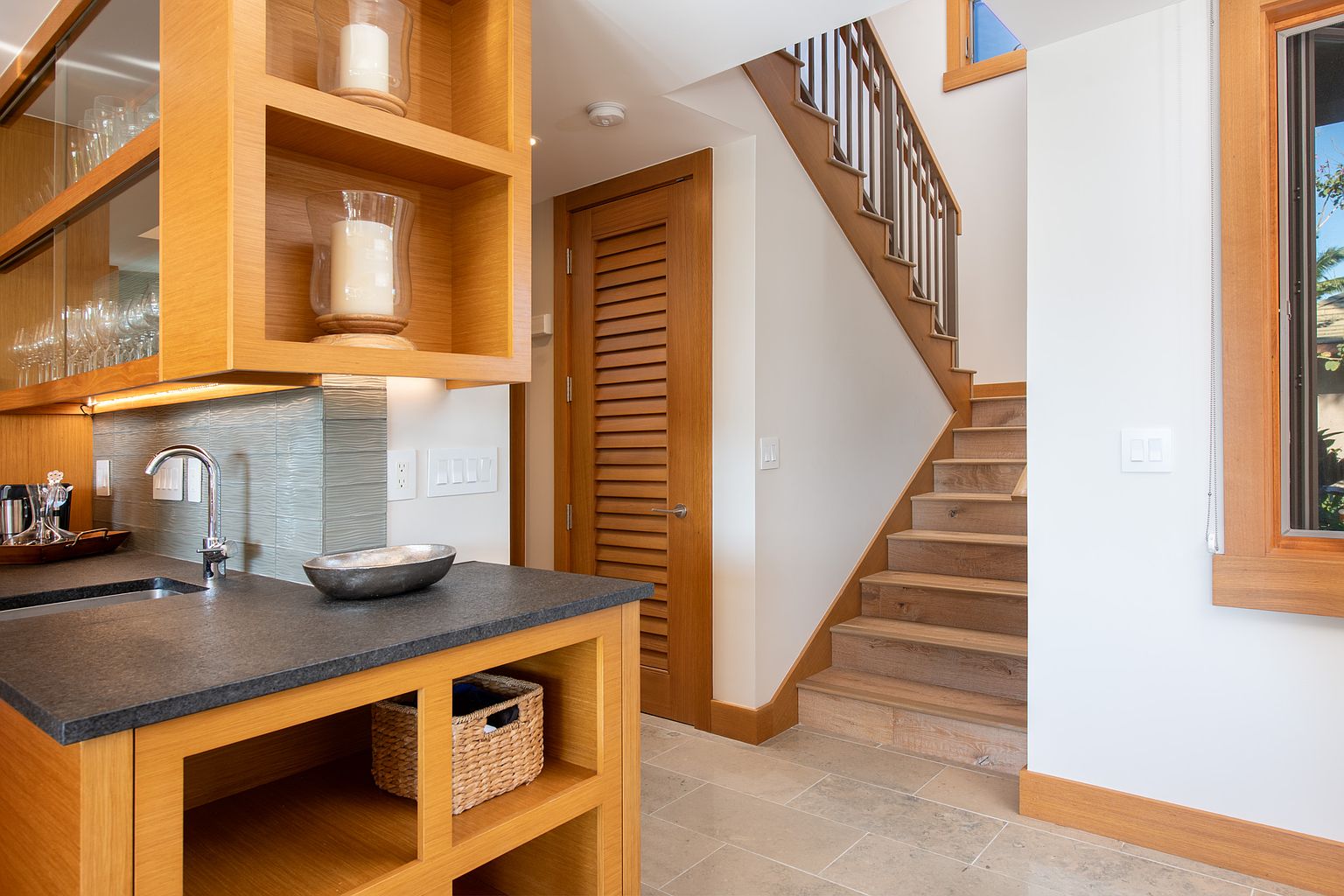 This interior shot showcases a hallway with wooden stairs and built-in cabinetry. The stairs feature a modern metal railing and light wood treads, while the cabinetry includes open shelving and a dark countertop with a sink. A louvered door is visible, adding texture and visual interest to the space.