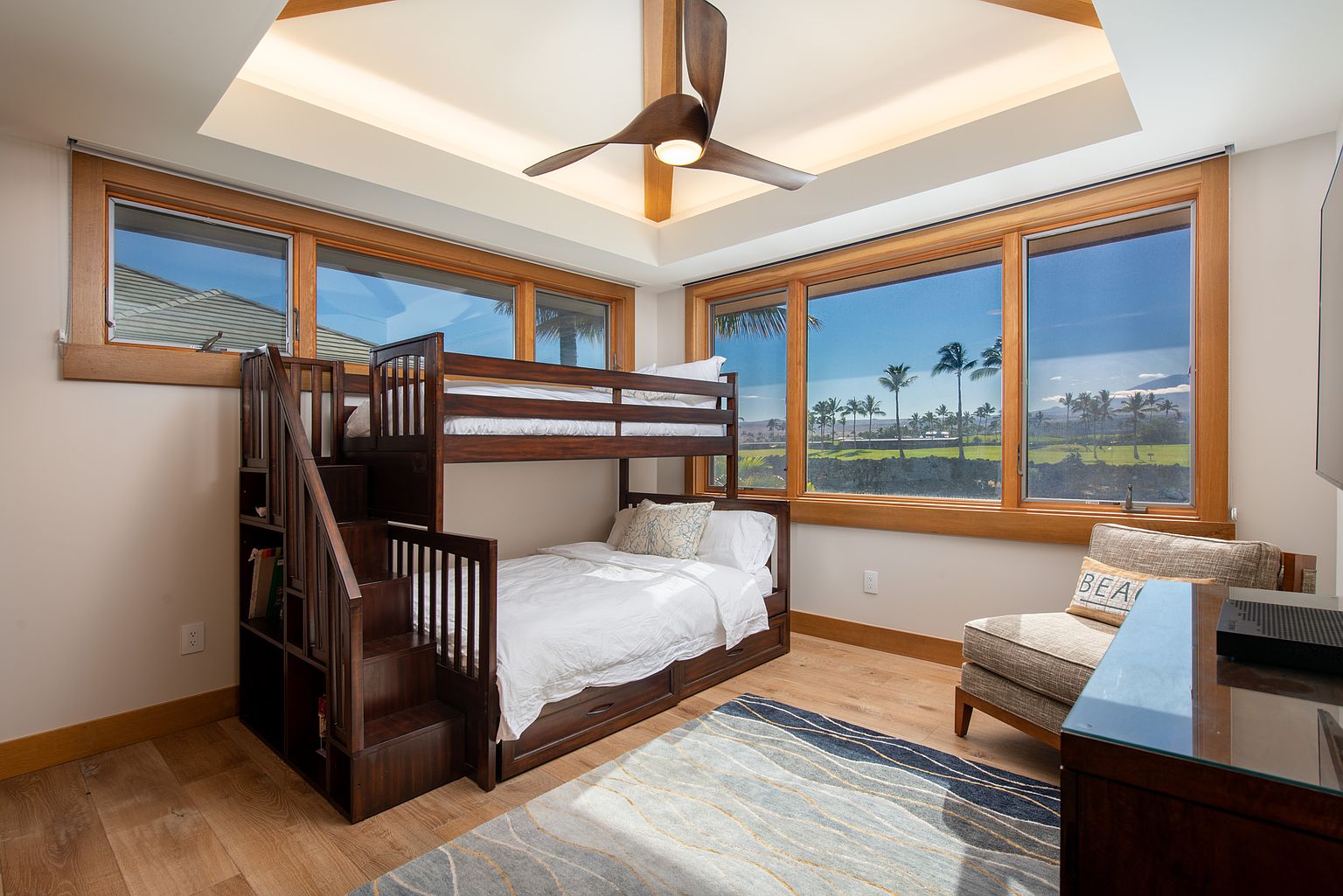 This is a cozy bedroom featuring a wooden bunk bed with built-in stairs and storage. Large windows offer a view of palm trees and the ocean, creating a relaxing atmosphere. The room is decorated with a neutral color palette and a patterned rug, enhancing the inviting feel.