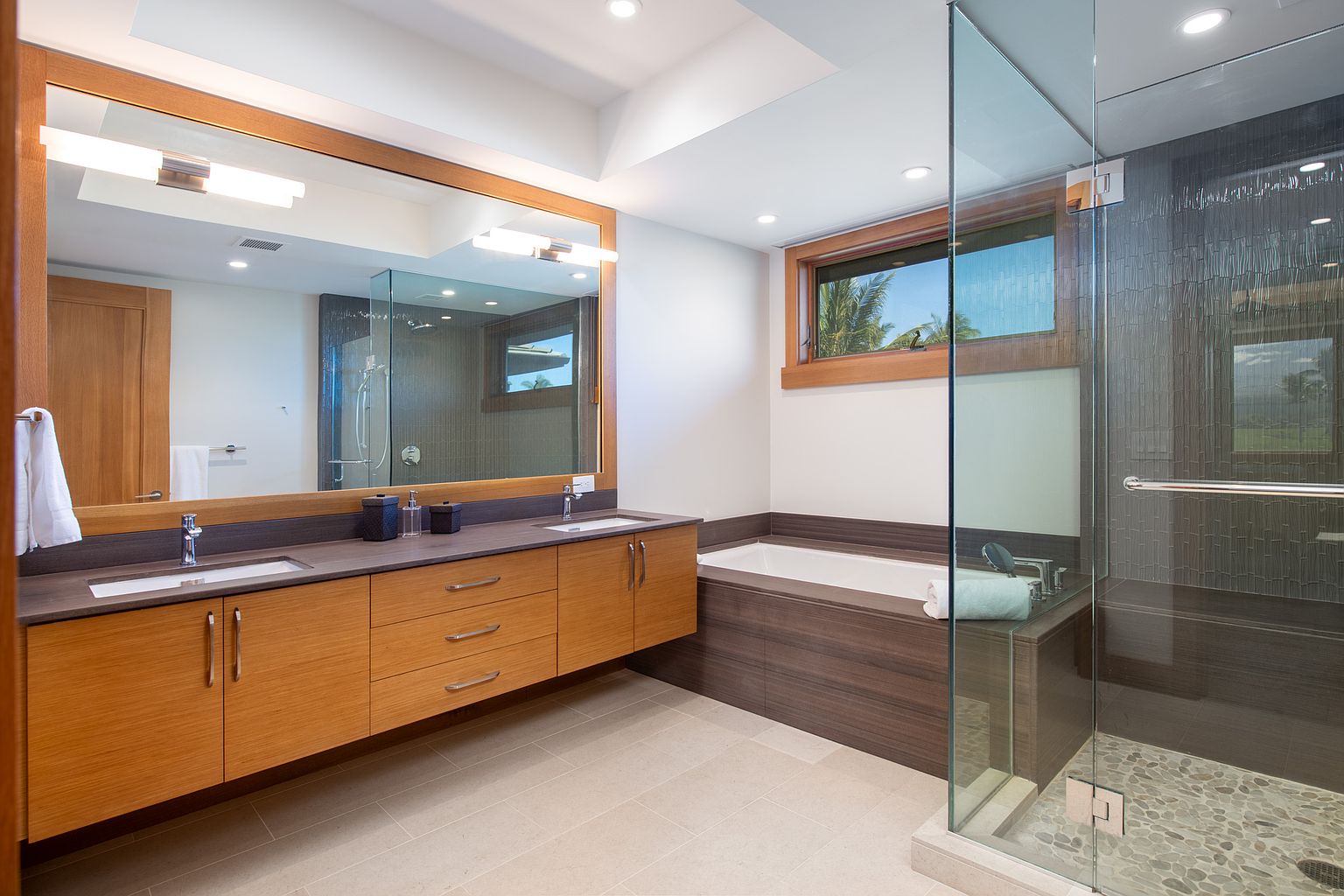This is a modern primary bathroom featuring a double vanity with wood cabinetry and a dark countertop. A large mirror spans the length of the vanity, reflecting the natural light. The bathroom also includes a glass-enclosed shower and a separate bathtub, creating a spa-like atmosphere.