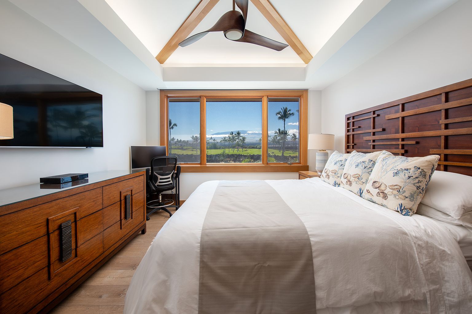 This is a primary bedroom featuring a king-size bed with a white duvet and decorative pillows, a wooden headboard, and a dresser with a large flat-screen TV. The room has a modern ceiling fan and a large window with a view of a golf course and palm trees. The overall style is luxurious and comfortable.
