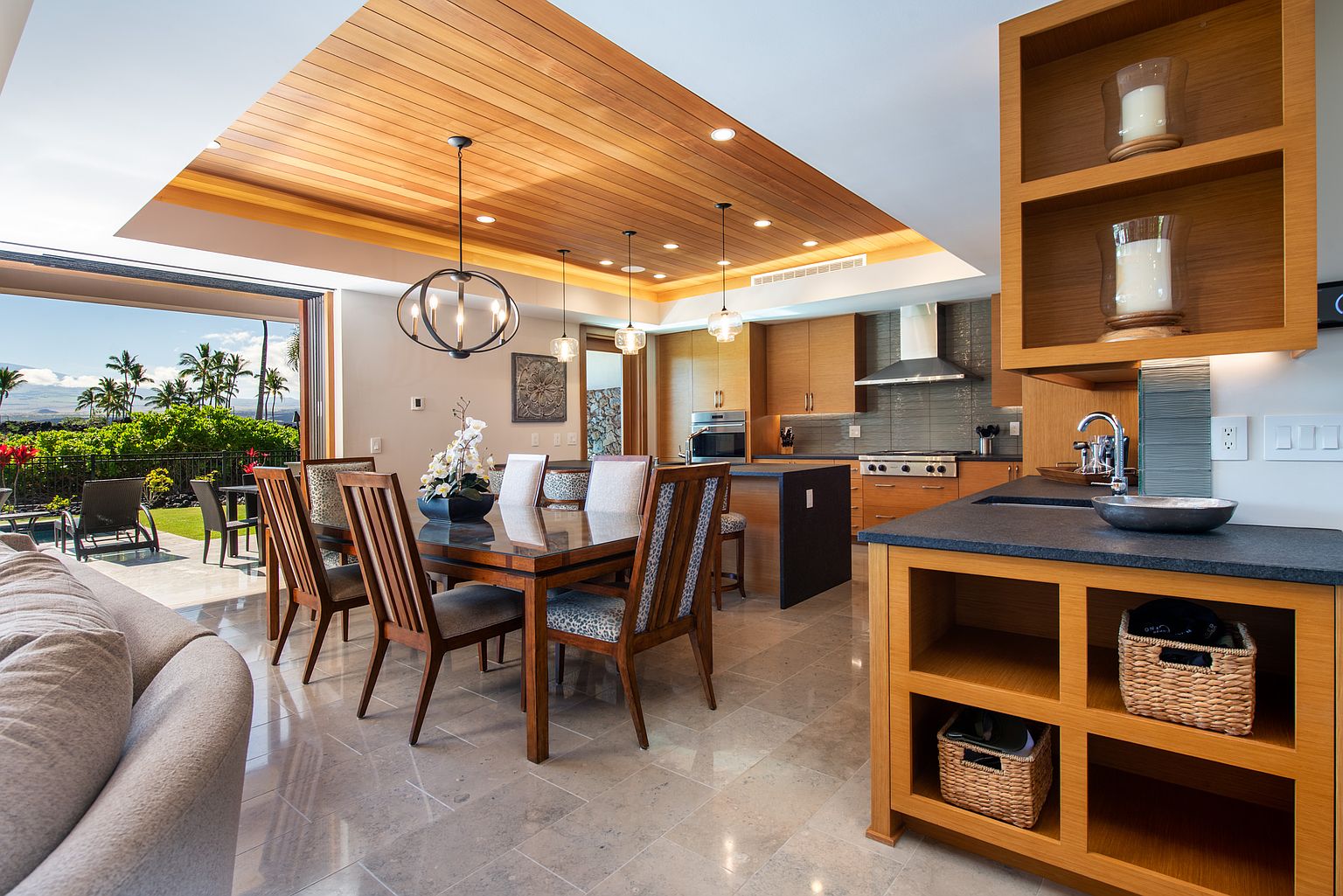This interior shot showcases a dining room seamlessly connected to the kitchen and an outdoor patio. The dining area features a wooden table with seating for six, complemented by modern lighting fixtures and a wooden ceiling. The open layout enhances the sense of space and luxury, perfect for entertaining.