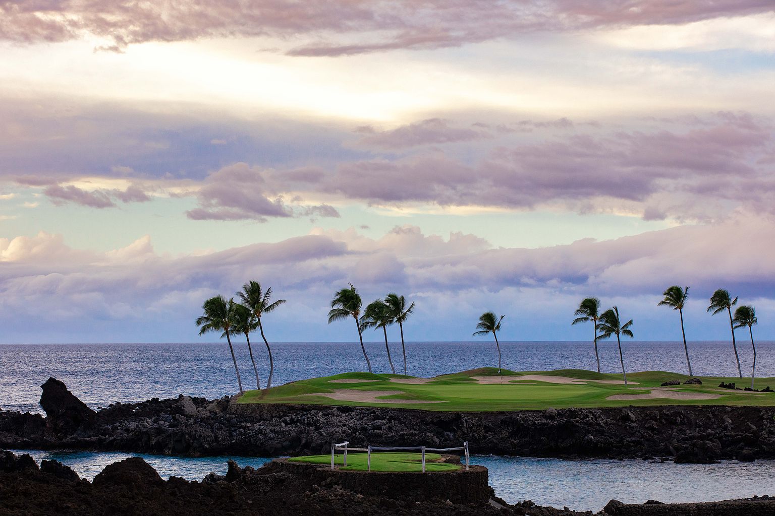 This scenic shot showcases a lush green golf course extending to the ocean, framed by dramatic lava rock formations. Palm trees dot the landscape, adding a tropical touch under a sky filled with soft, pastel-colored clouds. The image evokes a sense of luxury and tranquility, perfect for highlighting the property's access to exclusive amenities and stunning natural surroundings.