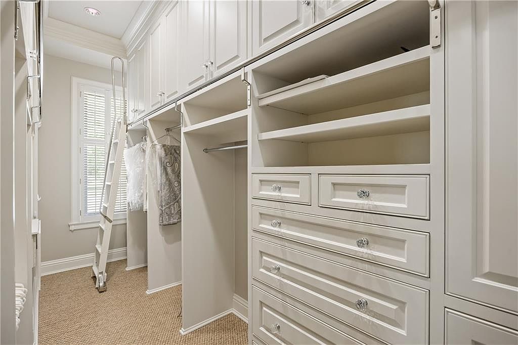 This spacious walk-in closet features custom light-colored cabinetry with extensive shelving, hanging rods, and a series of drawers with decorative crystal-style knobs. A rolling library-style ladder is positioned against the wall, providing access to the high-reach storage compartments above. The room is finished with neutral wall tones and textured carpeting, creating a clean and organized aesthetic.