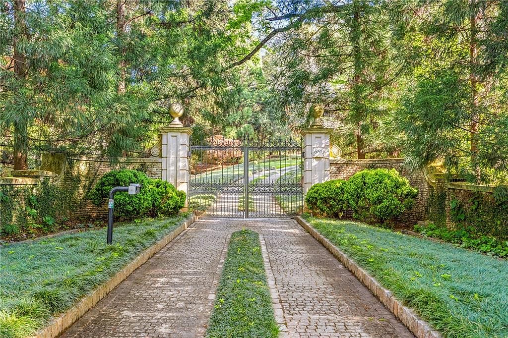 This image captures a grand, gated entrance to a private estate, featuring a cobblestone driveway flanked by lush, manicured greenery and mature trees. The focal point is a pair of ornate black wrought-iron gates set between two substantial stone pillars topped with decorative spheres. The perspective is a straight-on, eye-level shot that emphasizes the sense of privacy and exclusivity of the property.
