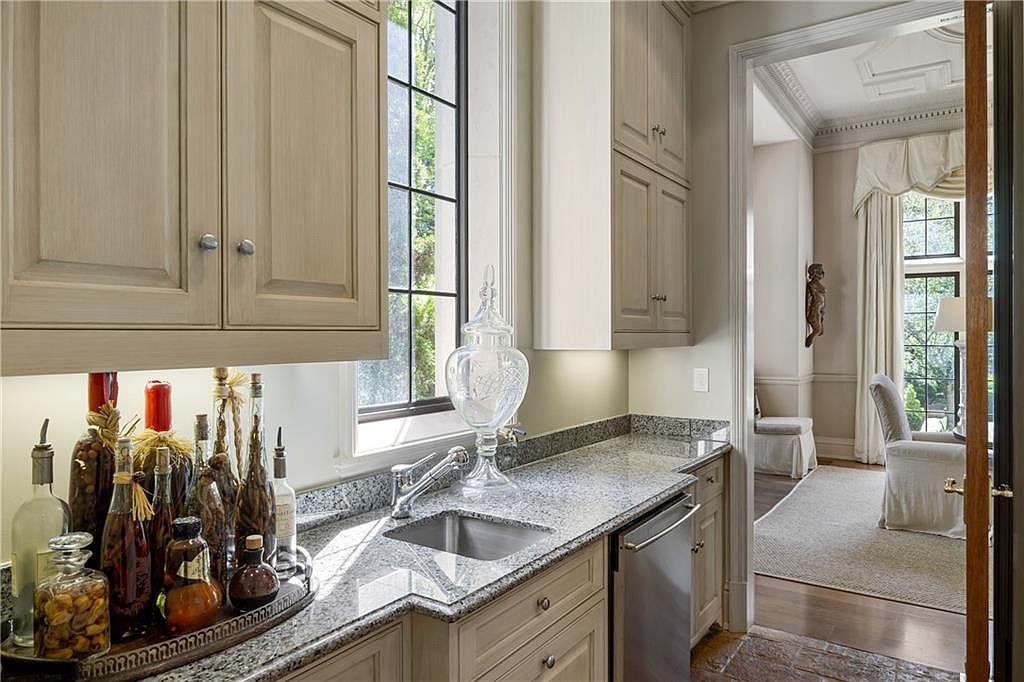 This elegant wet bar or butler's pantry features light-toned cabinetry, speckled stone countertops, and a stainless steel under-mount sink. A collection of decorative glass bottles and a large crystal decanter sit on the counter, while a doorway leads into a bright, formal living area with tall windows and draped curtains. The perspective captures the transition between the functional service area and the refined living space, highlighting the home's sophisticated design.