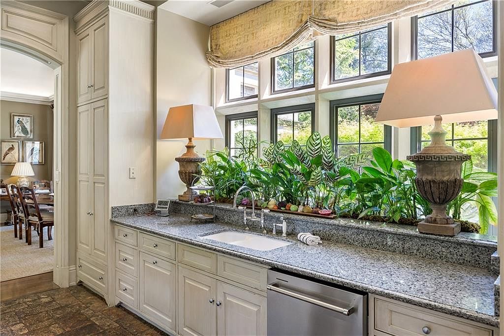This elegant kitchen features light-toned cabinetry and speckled stone countertops, centered around a bright sink area positioned beneath a large window bank. Lush greenery sits on the windowsill, flanked by two decorative table lamps that add a warm, sophisticated touch to the workspace. The perspective captures the functional layout of the kitchen while offering a glimpse into an adjacent dining area through an arched doorway.