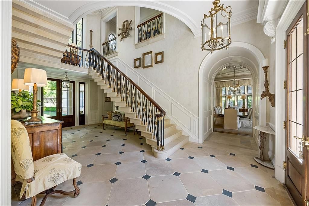 This grand foyer features a sweeping staircase with an ornate metal railing and stone steps, serving as the focal point of the space. The area is finished with light-toned stone flooring accented by dark square inlays, leading toward an arched entryway that reveals a formal dining room. A large, multi-tiered brass chandelier hangs from the ceiling, casting a warm glow over the elegant, traditional architectural details.