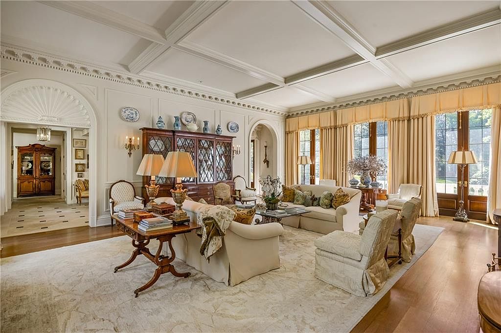 This elegant, formal living room features a coffered ceiling, intricate crown molding, and a neutral color palette that creates a sophisticated atmosphere. The space is furnished with plush seating, a central coffee table, and a large wooden hutch, all arranged on a patterned area rug over hardwood floors. Tall windows draped in floor-length curtains allow natural light to fill the room, while an arched doorway provides a view into an adjacent space with a secondary cabinet.