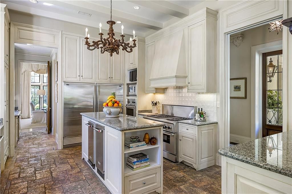 This elegant kitchen features cream-colored cabinetry, a central island with a stone countertop, and stainless steel appliances, including a large refrigerator and a professional-style range. The space is anchored by a decorative chandelier and finished with rustic stone flooring, creating a warm and sophisticated atmosphere. The perspective looks directly into the kitchen, showcasing the functional layout and the transition to an adjacent room through a wide doorway.