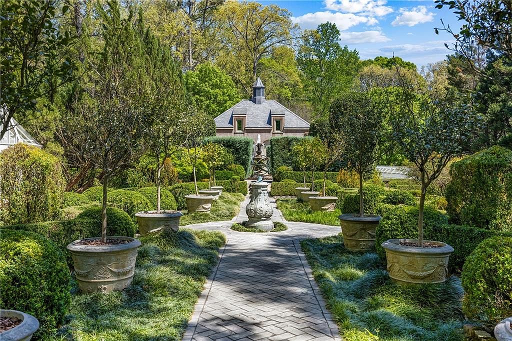 This meticulously manicured formal garden features a central stone-paved path leading toward a classic stone structure with a slate roof. The path is flanked by symmetrical rows of potted trees and neatly trimmed boxwood hedges, creating a structured and elegant landscape design. A decorative stone armillary sphere serves as a focal point in the center of the walkway, enhancing the sophisticated, estate-like atmosphere.