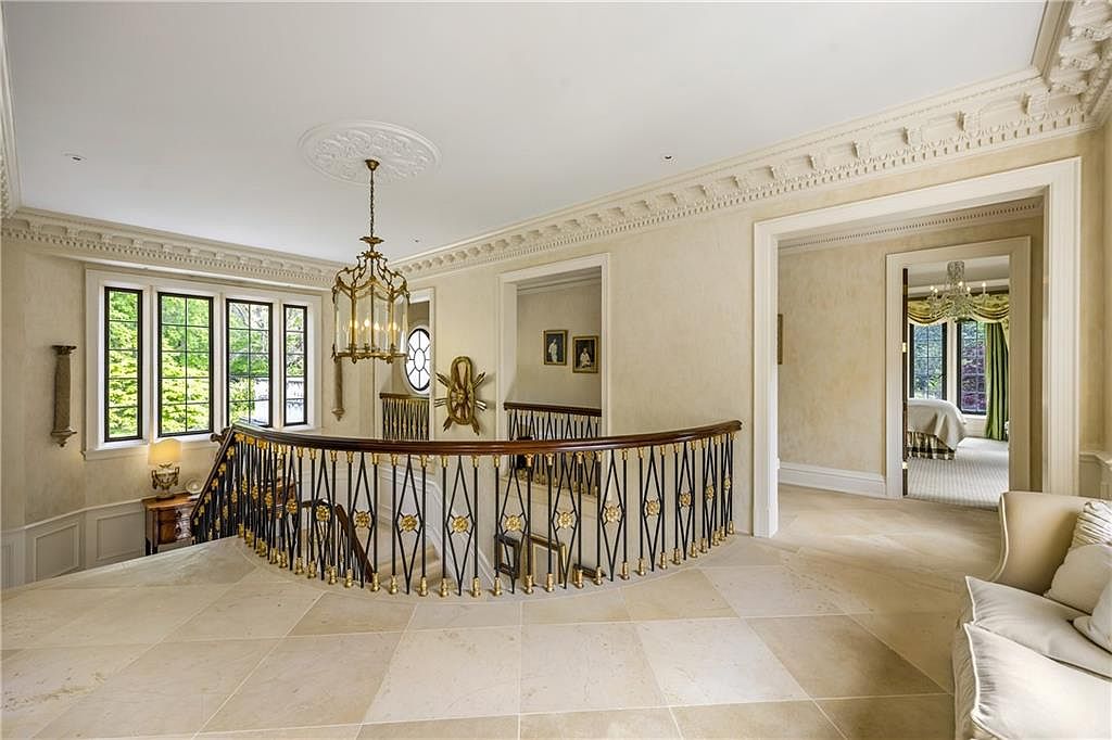 This elegant landing area features a curved staircase with ornate metal railings and a polished wood handrail, overlooking the level below. The space is finished with light-toned stone flooring, intricate crown molding, and a classic chandelier that serves as a focal point. A doorway leads into a bright bedroom, while the neutral color palette and architectural details create a sophisticated and airy atmosphere.
