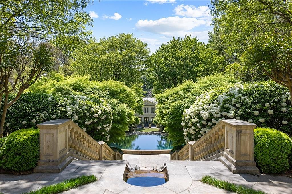 This elegant garden scene features a grand stone staircase leading down toward a serene, circular swimming pool nestled among lush greenery. The perspective is captured from the top of the stairs, looking down through a frame of mature trees and blooming white hydrangea bushes toward a classic residence in the distance. The composition emphasizes a formal, symmetrical landscape design that evokes a sense of luxury and tranquility.