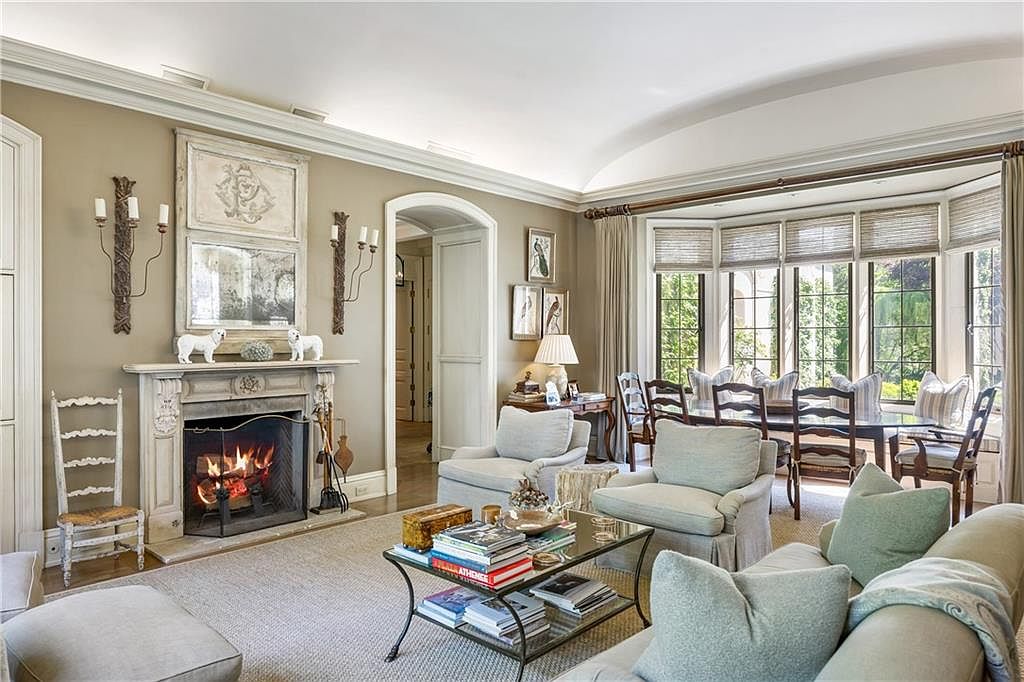 This elegant living room features a neutral color palette, a classic fireplace with a decorative mantel, and comfortable seating arranged around a glass-topped coffee table. Large windows with woven shades allow natural light to fill the space, while an adjacent dining area is visible through the open layout. The room exudes a sophisticated and inviting atmosphere, perfect for both relaxation and formal entertaining.