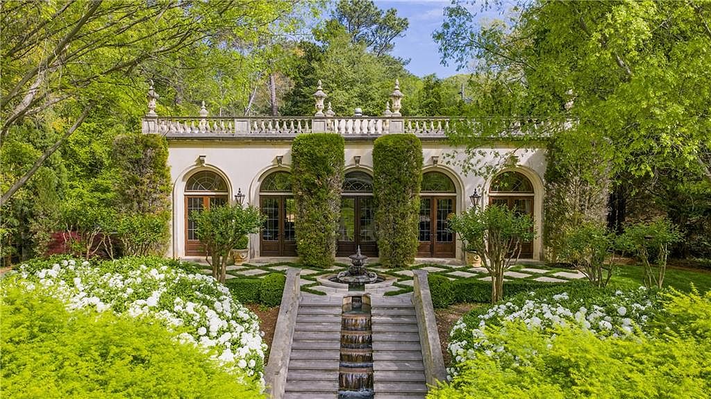 This elegant garden scene features a classical stone structure with arched glass doors and a balustrade roofline, set against a backdrop of lush green trees. A tiered stone staircase with a central water feature leads up to the building, flanked by manicured hedges and vibrant white flowering shrubs. The perspective is a straight-on, eye-level shot that emphasizes the symmetrical, formal landscaping and the sophisticated architectural design.
